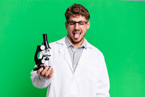 young adult caucasian man with cheerful and rebellious attitude, joking and sticking tongue out. scients laboratory student with a microscope concept - Fotografie, Obrázek