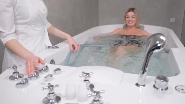 Una massaggiatrice massaggia una ragazza in una vasca da bagno con una corrente d'acqua. Giovane donna durante un idromassaggio in un salone di bellezza. Concetto di cura del corpo e trattamenti termali - Filmati, video