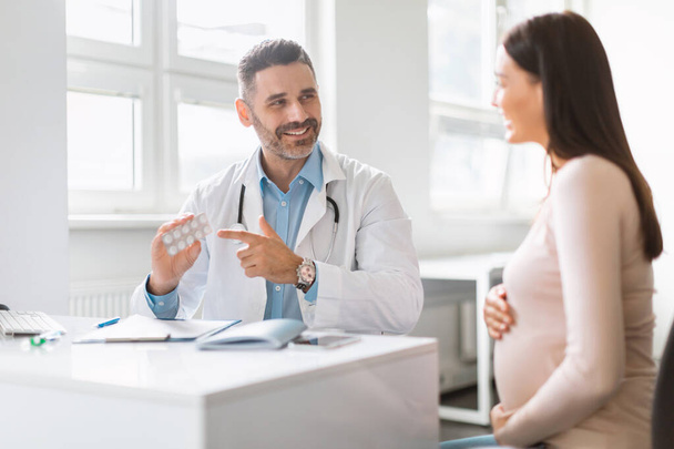 Healthy habits for pregnancy. Happy male therapist prescribing vitamins for young pregnant woman, pointing at blaster with pills, giving tablets to expectant mother during appointment in office - Foto, Imagen