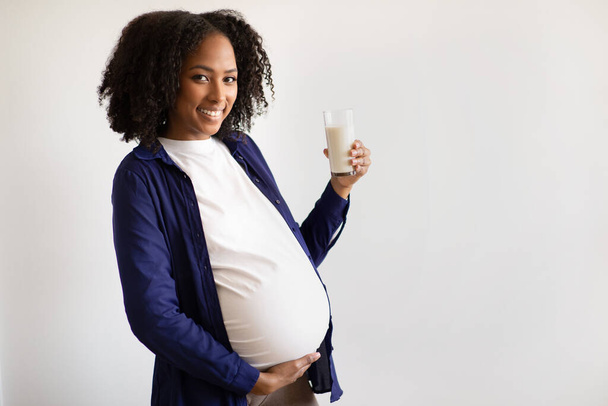 Веселая африканская беременная женщина тысячелетия с большим животом держит стакан молока на сером фоне стен. Ожидание ребенка, здравоохранение, диета на дому, объявление и предложение - Фото, изображение