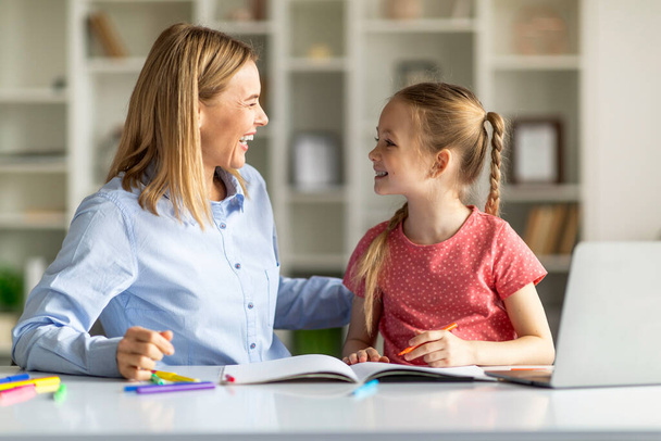 Mother And Her Cute Little Daughter Having Fun While Making School Homework Together, Happy Mom And Female Child Sitting At Desk In Home Interior And Smiling To Each Other, Closeup Shot - Foto, Bild