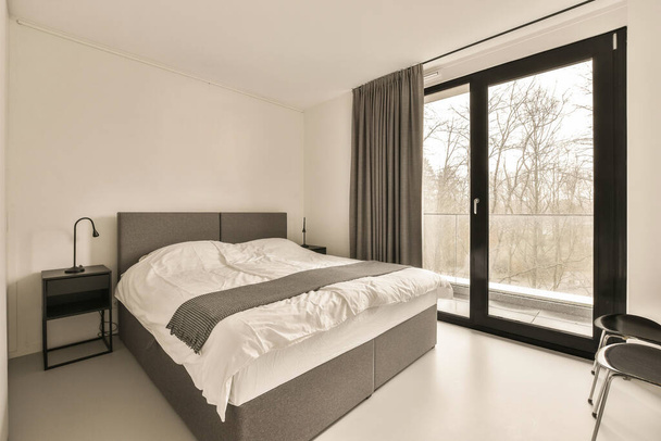 a bedroom with a bed, desk and large window looking out onto the trees in the photo is black and white - Foto, Imagem