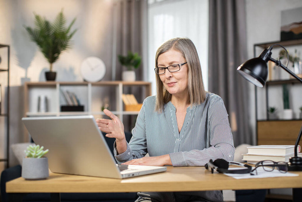 Beautiful female of mature age making video call via laptop camera while working at writing desk in home interior. Confident company leader negotiating contract online via internet application. - 写真・画像