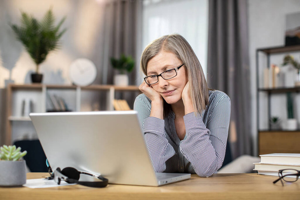 Exhausted middle-aged woman in glasses feeling tired while looking at laptop screen after full day of work in remote office. Senior lady being overcome with fatigue after online brainstorm session. - 写真・画像