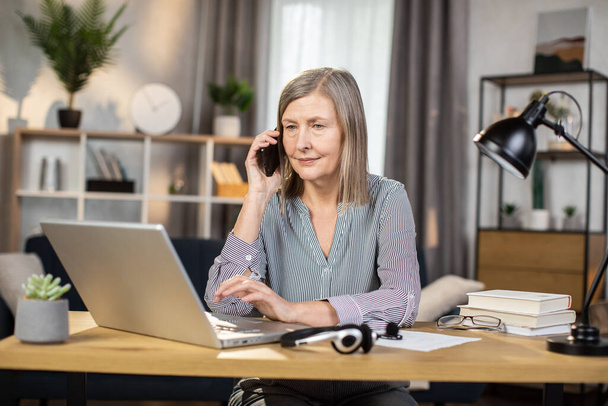 Thoughtful senior lady speaking on smartphone while using laptop touchpad placed on wooden desk of remote office. Experienced specialist having business conversation in company designated worksite. - Фото, зображення