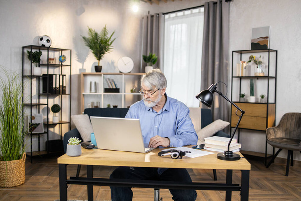 Focused mature adult in formal clothes typing on laptop while taking seat at modern desk in distant workplace. Respectful businessman in glasses checking email account on company website from home. - Foto, Bild