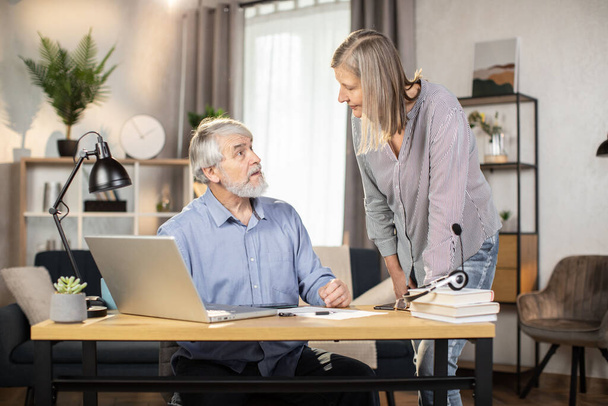 Mature couple of business people talking near writing desk with office equipment and stationery at home. Friendly spouses being involved in discussion of company matters in distant workplace. - Фото, изображение