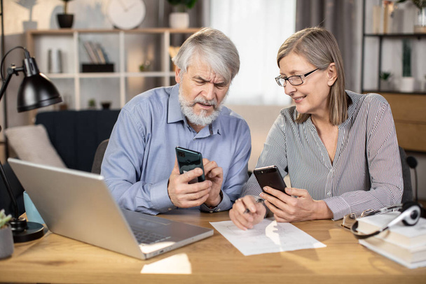 Close up view of mature businesspeople with cell phones looking at each other while staying in comfortable workplace. Senior husband and wife running business together using modern technologies. - Photo, Image