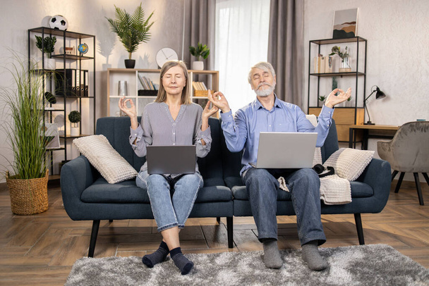 Full length view of calm mature male and female with computers sitting on cozy sofa with hands in gyan mudra. Senior adults relieving stress after hard working day doing yoga posture at home. - Photo, image