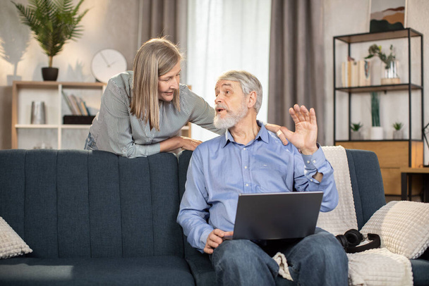 Beautiful mature lady helping senior husband with video call app on laptop while standing behind couch in living room. Elderly adults starting conversation with grown-up children via online app. - Photo, image