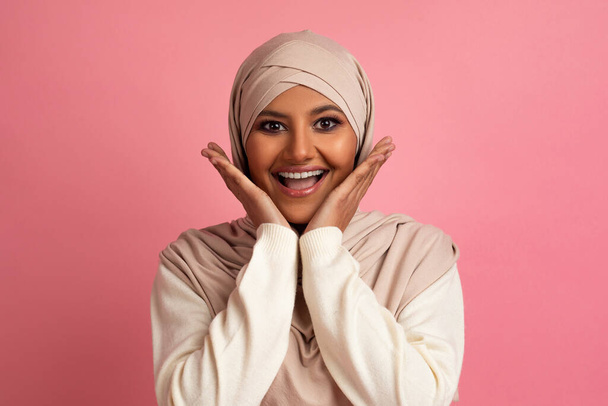 Große Überraschung. Fröhliche Muslimin, die ihr Gesicht berührt und aufgeregt ausruft, fröhliche junge islamische Frau, die emotional auf gute Nachrichten oder Angebote reagiert, posiert auf rosa Hintergrund, Kopierraum - Foto, Bild