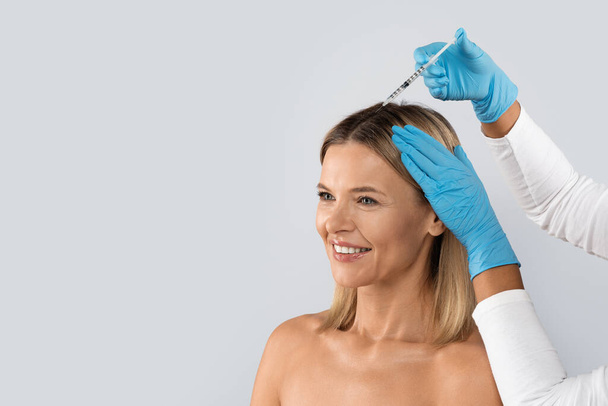 Hair loss, alopecia treatment. Unrecognizable beautician wearing medical gloves holding syringe, making scalp injections for smiling middle aged pretty blonde lady, copy space, studio background - Photo, Image
