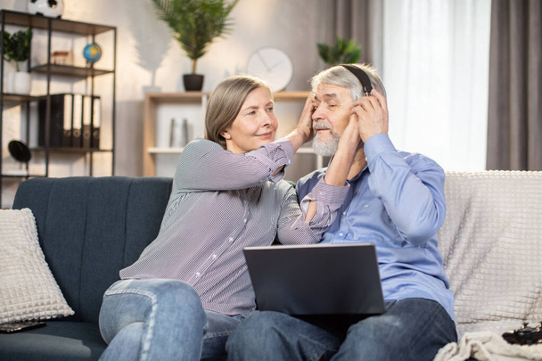 Lovely elder married couple resting on couch of beautiful apartment and using personal laptop for watching romantic movies. Caring wife helping husband to put on wireless headphones. - Valokuva, kuva