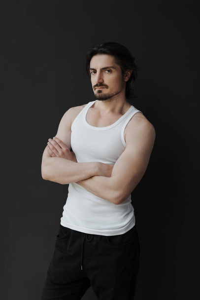 Self assured muscular bearded man with black hair in white sleeveless t shirt and pants standing on black background and looking at camera - Foto, Imagem