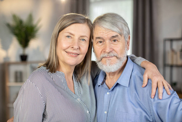Portrait of devoted couple standing close together, embracing and looking straight at camera. Mature woman holding hand gently on shoulder of grey-haired husband. Concept of marital love and care. - Photo, Image