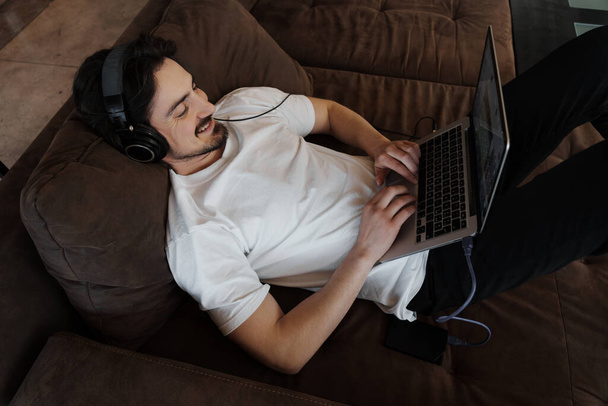 From above of smiling man in white t shirt and headphones composing music via laptop while lying on sofa - Fotografie, Obrázek