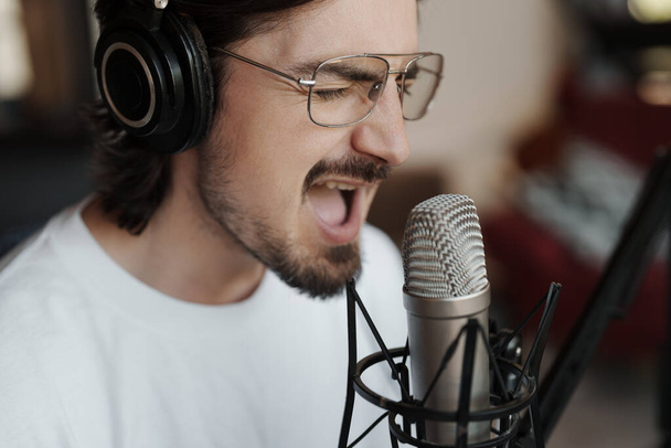 Вид збоку пристрасного співака-чоловіка в навушниках, що сидить за столом біля мікрофона і записує пісню в сучасній студії
 - Фото, зображення