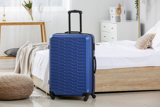 薄暗い部屋のベッドの近くの青いスーツケース - 写真・画像
