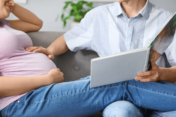 Young pregnant couple reading book on sofa at home, closeup - Фото, изображение