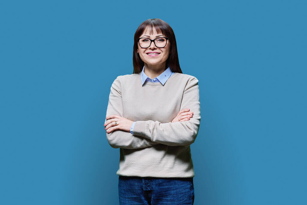 Positive middle aged woman posing over blue studio background. Mature smiling female in glasses with crossed arms looking at camera. Age, profession, job, work, service, business people concept - Fotó, kép