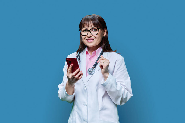 Mature female doctor in white lab coat using phone, on blue studio background. Medicine, online internet service, occupation, healthcare, health care, professional mobile apps, treatment concept - Фото, изображение