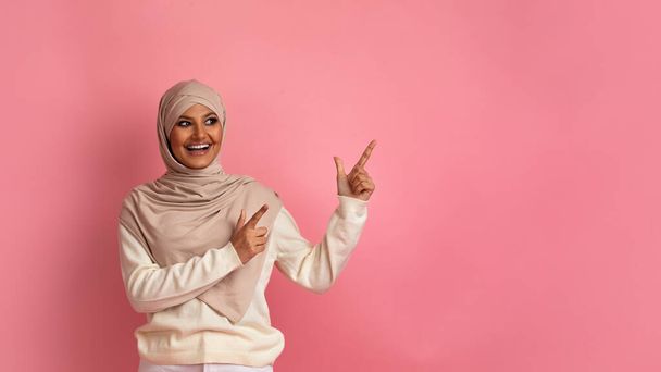 Special Offer. Cheerful Muslim Lady In Hijab Pointing Aside At Copy Space With Two Fingers, Excited Happy Islamic Woman Demonstrating Free Place For Advertisement On Pink Studio Background - Foto, Imagem