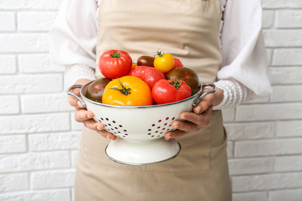 Woman holding colander with different fresh tomatoes on white brick background, closeup - Zdjęcie, obraz