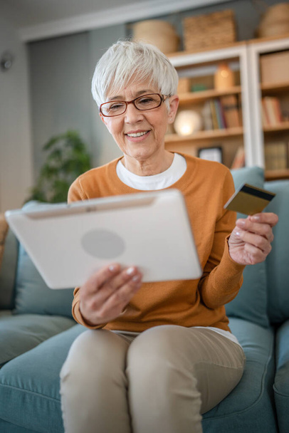 Yetişkin bir yaşlı kadın büyükanne evde oturup online alışveriş siteleri için kredi kartı ya da banka kartı kullanıyor. İndirimli indirimli ürünler alıyor. Gerçek insanlar dijital tablet kullanıyor. - Fotoğraf, Görsel