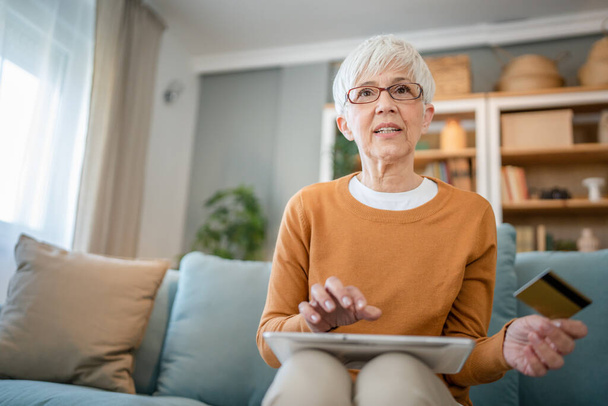 Egy érett idős nő nagymama ül otthon hitel- vagy betéti kártya online vásárlás böngészés internetes áruházak vásárol dolgokat akciós áron valódi emberek másolni tér használata digitális tabletta - Fotó, kép
