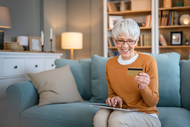 Одна зрелая пожилая женщина бабушка сидеть дома использовать кредитную или дебетовую карту для интернет-магазинов просматривать покупки вещи на скидку продажи реальные люди копировать пространство использовать цифровые планшеты - Фото, изображение