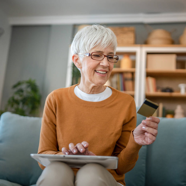 Yetişkin bir yaşlı kadın büyükanne evde oturup online alışveriş siteleri için kredi kartı ya da banka kartı kullanıyor. İndirimli indirimli ürünler alıyor. Gerçek insanlar dijital tablet kullanıyor. - Fotoğraf, Görsel
