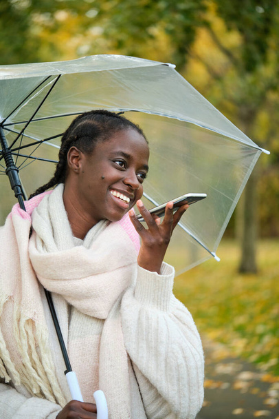Afrykanka z przezroczystym parasolem uśmiecha się i nagrywa wiadomość audio jesienią. - Zdjęcie, obraz