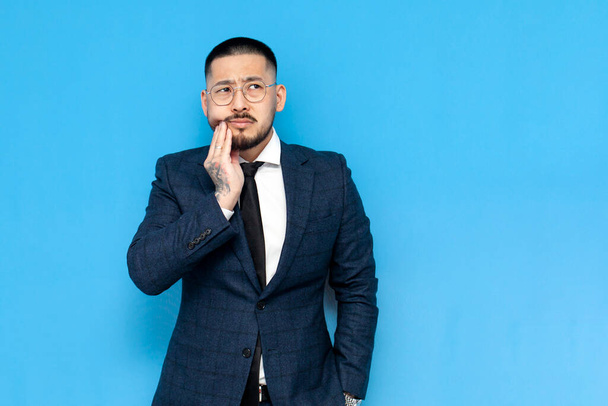 unglücklicher asiatischer Geschäftsmann in Anzug und Brille hält Zahn mit den Händen auf blauem Hintergrund und ist traurig, koreanischer Unternehmer in Businesskleidung zeigt Zahnschmerzen und blickt in den Kopierraum - Foto, Bild
