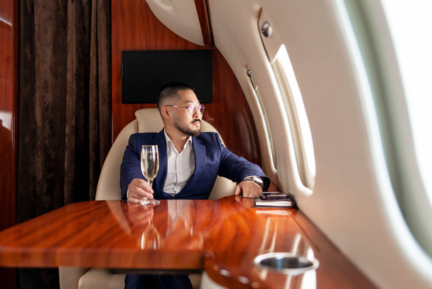 succès homme d'affaires asiatique en costume et verres avec verre de champagne mouches dans jet de luxe privé, entrepreneur coréen repos et détente en vol, style de vie de luxe - Photo, image