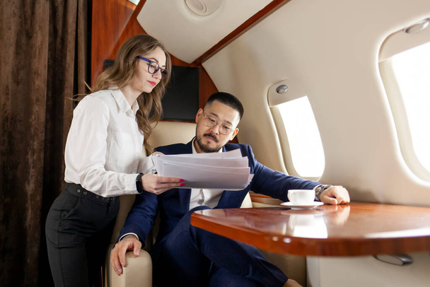 Takım elbiseli ve gözlüklü başarılı Asyalı işadamı, sekreter yardımcısıyla birlikte lüks bir jette uçuyor. Kadın yönetici, belgelerini ve raporlarını uçaktaki patrona gösteriyor. - Fotoğraf, Görsel