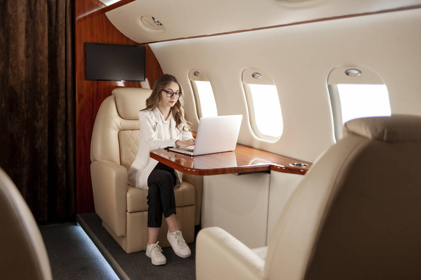 бізнес-леді в костюмі і окулярах летить в приватному розкішному літаку і працює на ноутбуці, менеджер дівчини фінансист використовує комп'ютер і типи в літаку
 - Фото, зображення