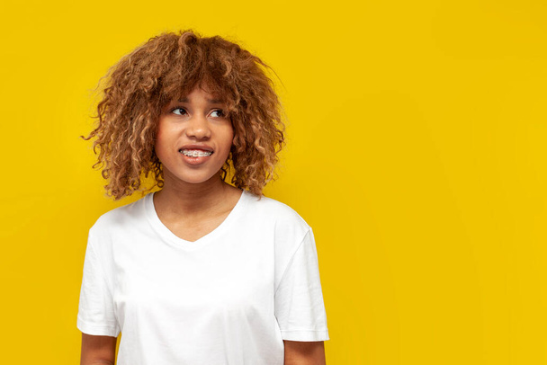 young satisfied curly american girl with braces bites her lip and dreams on yellow isolated background, african woman with hairstyle and toothy smile imagines and looks at the copy space - Fotografie, Obrázek