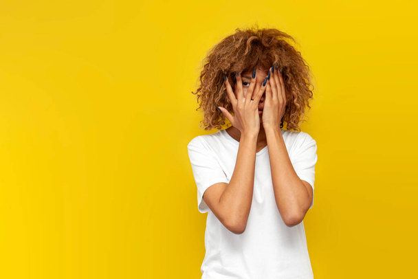 young curly american girl covers her face with her hands and peeps on yellow isolated background, african frightened woman in white t-shirt hides and looks at the copy space - Fotó, kép
