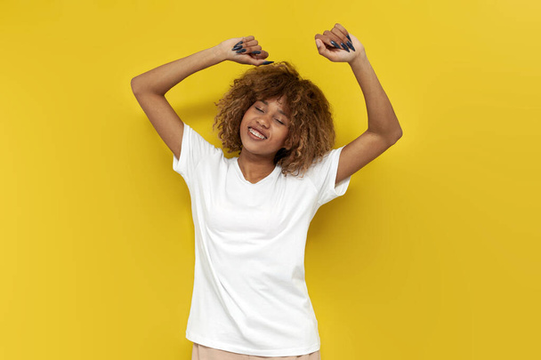 young curly american girl dancing to the music on yellow isolated background, african woman in white t-shirt with her eyes closed moves with her hands up - Photo, Image
