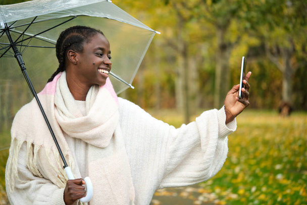 Afrikkalainen nainen ottaa selfie älypuhelimella ja läpinäkyvä sateenvarjo hymyilee syksyllä. - Valokuva, kuva