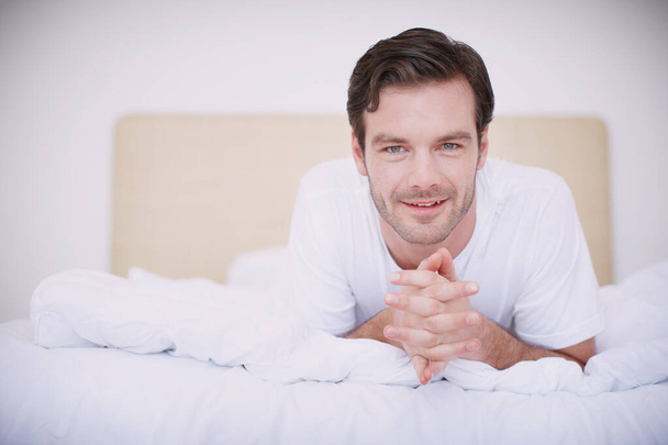 Расслабляюсь в постели. Портрет красивого молодого человека, лежащего на животе на кровати с сжатыми руками - Фото, изображение