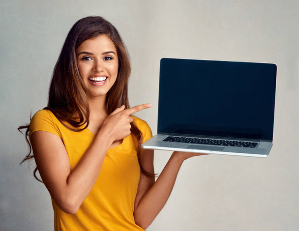 Este sitio web es simplemente maravilloso. Retrato de estudio de una joven atractiva apuntando a un portátil con una pantalla en blanco sobre un fondo gris - Foto, imagen