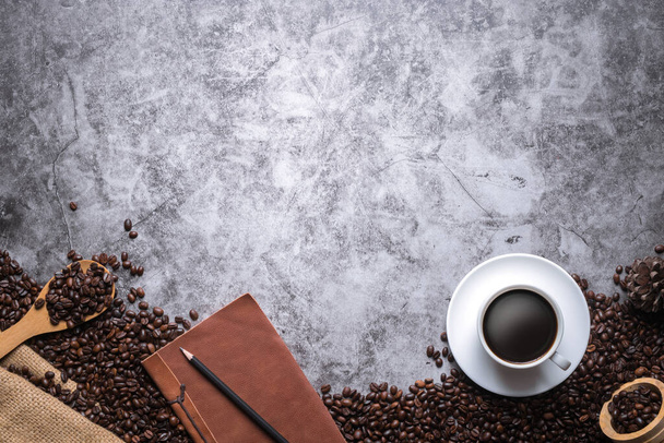 The coffee is in a white cup with a leather notebook and pencil placed laid on the old floor and surrounded by many coffee beans, with copy space. - Photo, Image