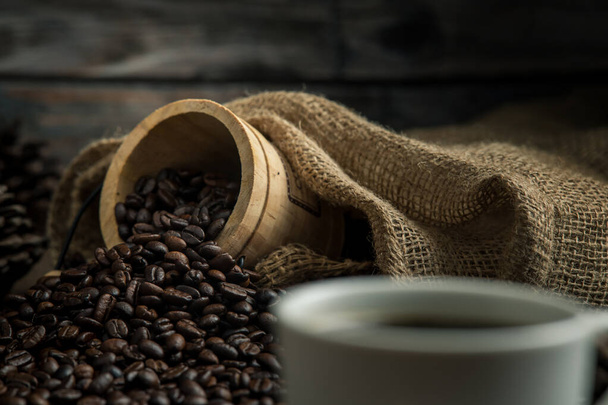 Gorąca kawa w białej filiżance kawy i zamknąć wiele ziaren kawy umieszczone wokół na drewnianym stole w ciepłej, jasnej atmosferze, na ciemnym tle - Zdjęcie, obraz