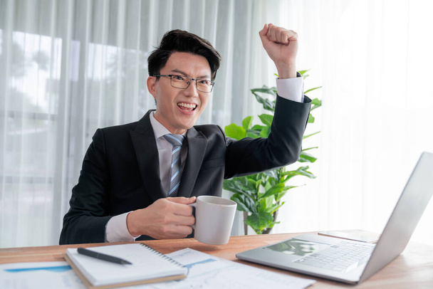 Aufgeregter asiatischer Geschäftsmann feiert Erfolg am Schreibtisch. Glücklicher Büroangestellter erreicht Geschäftsziele mit Datenanalyse oder Marketingplanung. Feierlicher Ausdruck des Geschäftssiegers. Jubelnd - Foto, Bild