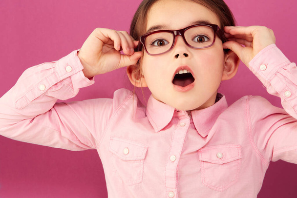 Portrait, shocked and girl child with glasses in studio isolated on a pink background. Kid, nerd and face of surprised person, wow or omg, emoji or reaction to unexpected news, announcement or secret. - Fotó, kép