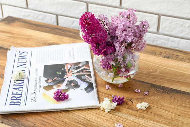 Glass vase of beautiful fragrant lilac flowers and magazine on wooden table near white brick wall - Photo, Image