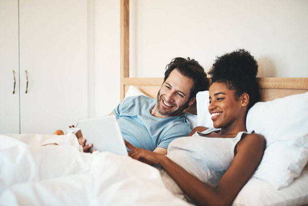 Happy couple, tablet and relax on bed for morning entertainment or online streaming together at home. Interracial man and woman person relaxing in bedroom on technology for social media or browsing. - Foto, Imagen