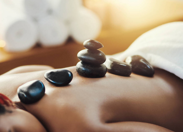 Woman, hot rocks and massage on back at spa for skincare, beauty or body treatment at resort. Calm female lying on bed with heated rock pile for healthy physical therapy, zen or wellness at salon. - Photo, Image