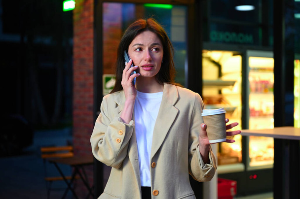 Una hermosa mujer vestida con atuendo casual está disfrutando de una taza de café en un entorno urbano. Ella parece estar profundamente en la conversación mientras habla por teléfono, con una expresión que sugiere que está enfocada y contenta. - Foto, imagen